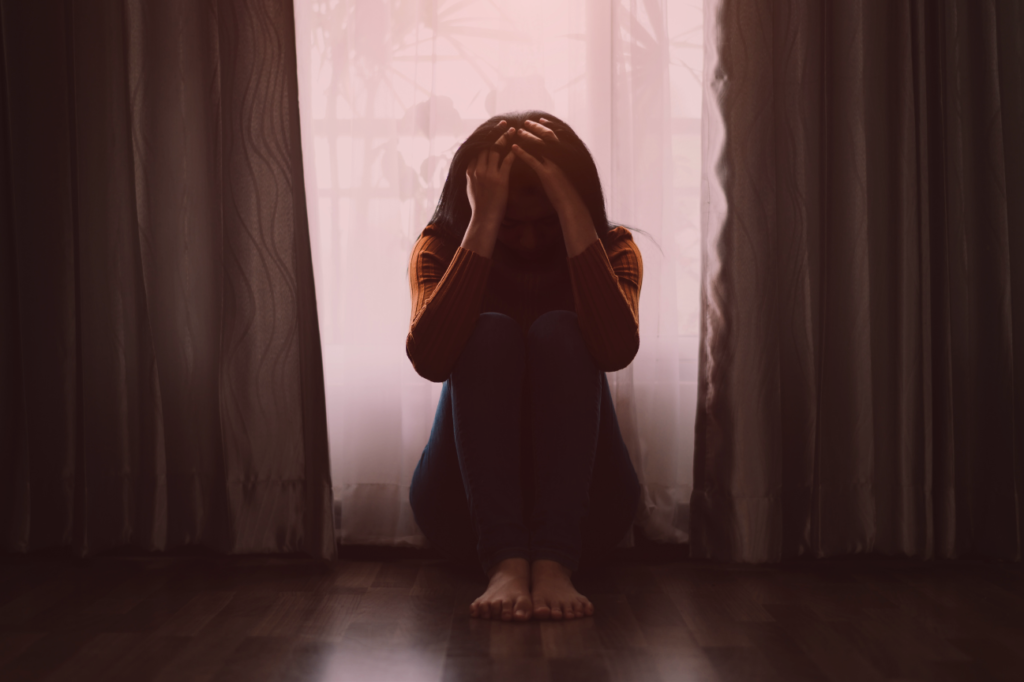 Pessoa sentada no chão com as mãos na cabeça, em posição de desespero e vulnerabilidade, em um ambiente escuro e silencioso, iluminada pela luz difusa de uma janela coberta por cortinas. Simbolizando que falsos pais de santo se aproveitam de pessoas vulneráveis.  