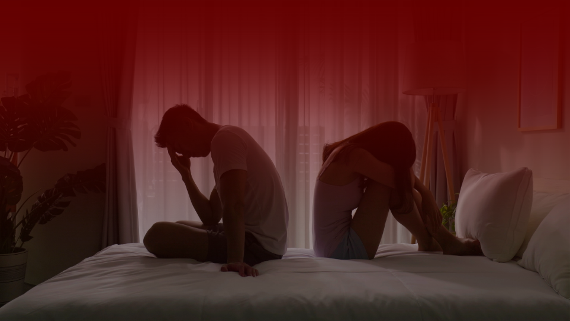 Imagem de um casal sentado na cama, ambos com expressões de preocupação, em um ambiente escuro iluminado suavemente pela luz da janela ao fundo, com tons de vermelho predominantes, simbolizando tensão emocional e necessidade de saber como melhorar seu relacionamento.