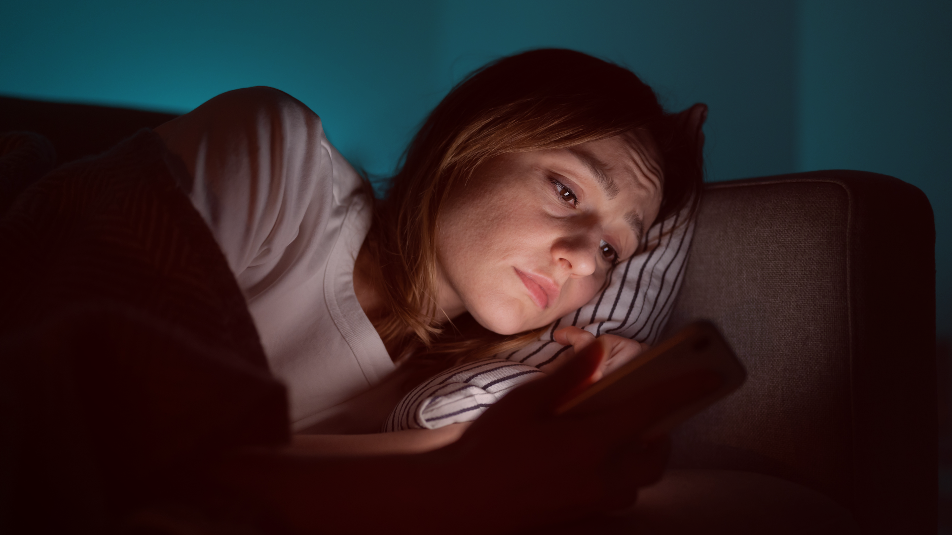 Mulher deitada no sofá, segurando um celular e olhando para a tela com expressão triste e preocupada. A iluminação suave e o ambiente escuro sugerem um momento de reflexão ou decepção,Simbolizando a ideia do artigo "ele te bloqueou".