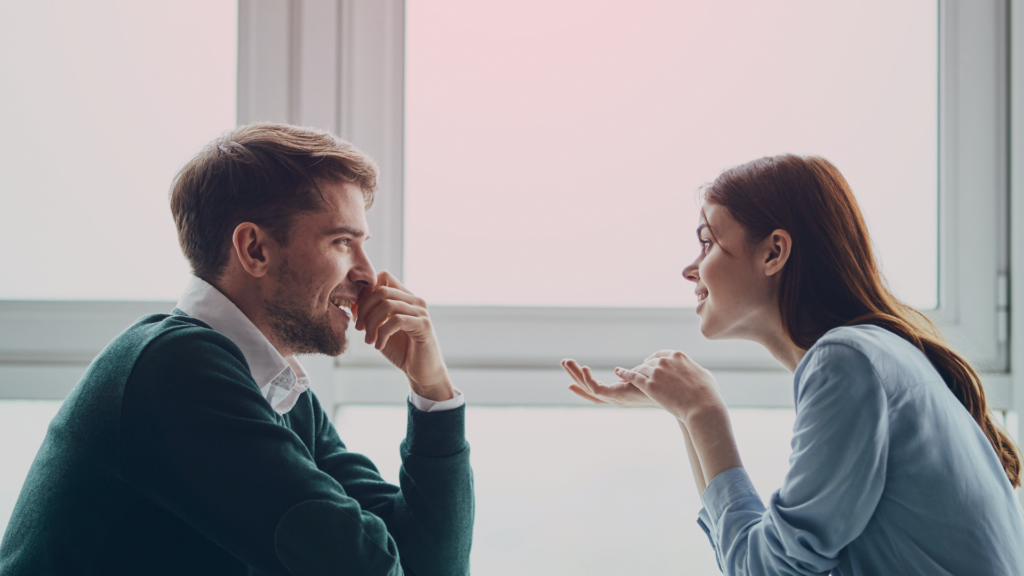 Homem e mulher sentados frente a frente, sorrindo e conversando animadamente. A mulher gesticula enquanto fala, e o homem escuta com atenção, demonstrando interesse e conexão. O fundo claro e a iluminação suave criam uma atmosfera acolhedora e harmoniosa.