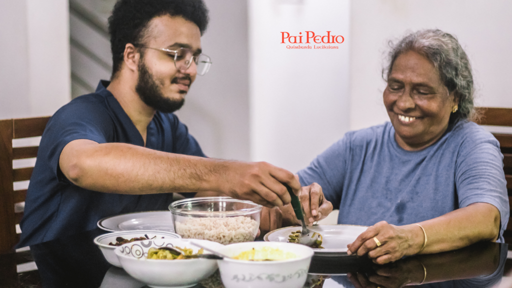 Cena calorosa e afetuosa de uma refeição em família. Um jovem homem serve comida com carinho no prato de uma senhora mais velha, que sorri com os olhos cheios de ternura. Ambos estão sentados à mesa, cercados por tigelas de alimentos caseiros. A atmosfera é de paz, reconciliação e cuidado mútuo. No topo da imagem, está o logotipo “Pai Pedro — Quimbanda Luciferiana”.