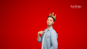 Homem com expressão de superioridade, usando uma coroa dourada e camisa jeans, em frente a um fundo vermelho. Imagem representa o comportamento típico de uma pessoa narcisista no relacionamento.