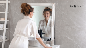 Imagem de uma mulher de roupão branco encarando seu reflexo no espelho e sorrindo, em um banheiro com design moderno e tons neutros. O ambiente transmite leveza, autocuidado e autoestima. No canto superior direito, o logotipo “Pai Pedro – Quimbanda Luciferiana” aparece discretamente, conectando o conceito de se valorizar com a energia espiritual de empoderamento feminino.