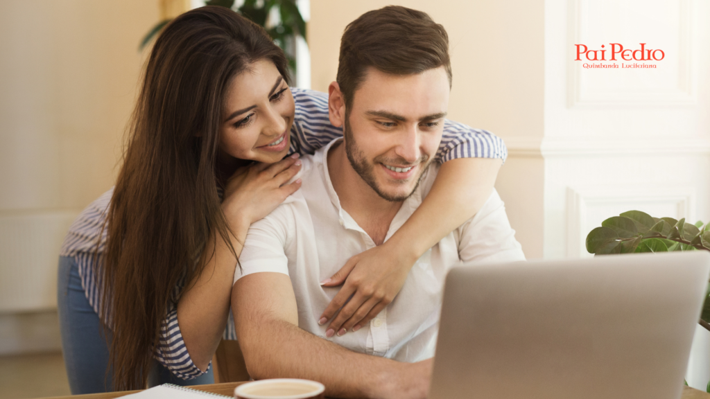 Casal sorrindo enquanto usa um notebook, com a mulher abraçando o parceiro por trás em um gesto carinhoso. A cena ilustra como surpreender namorado com atitudes simples, mostrando conexão, leveza e presença no dia a dia.