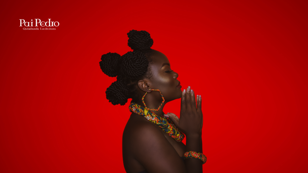 Mulher de perfil com pele negra e cabelo trançado em coque artístico, olhos fechados e mãos em posição de prece, diante de um fundo vermelho intenso. Ela usa acessórios e tecidos africanos coloridos, transmitindo serenidade, fé e ancestralidade.