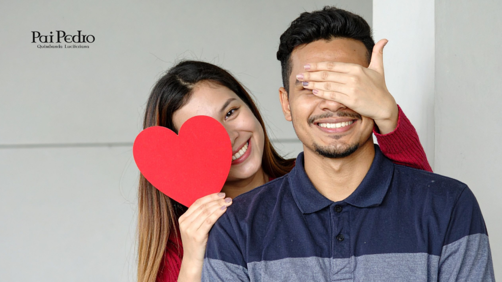 Casal sorrindo enquanto a mulher cobre os olhos do parceiro e segura um coração vermelho. A cena representa como surpreender namorado com gestos simples e inesperados que quebram a rotina e fortalecem o vínculo.