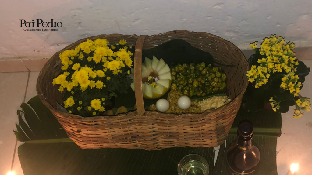 Cesta ritual adornada com flores amarelas, frutas, alimentos e elementos simbólicos dispostos sobre folhas verdes, acompanhada por velas e garrafa de bebida ritualística. A imagem representa uma oferenda preparada no Terreiro Pai Pedro da Quimbanda, em um ambiente de devoção e respeito às entidades espirituais.