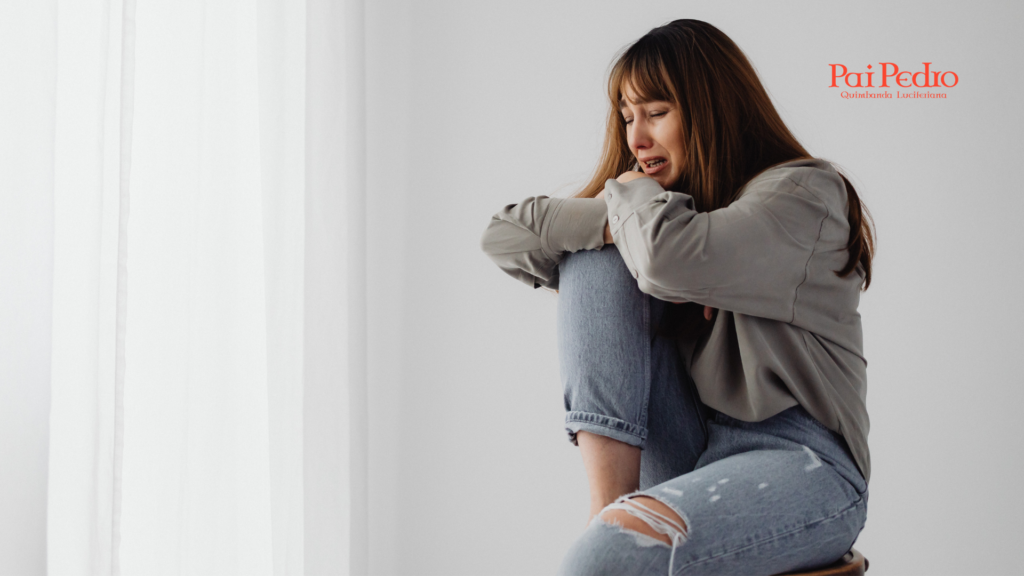 Mulher chorando sentada e abraçando as pernas em um ambiente claro e vazio, simbolizando dor emocional e os efeitos da solidão crônica.