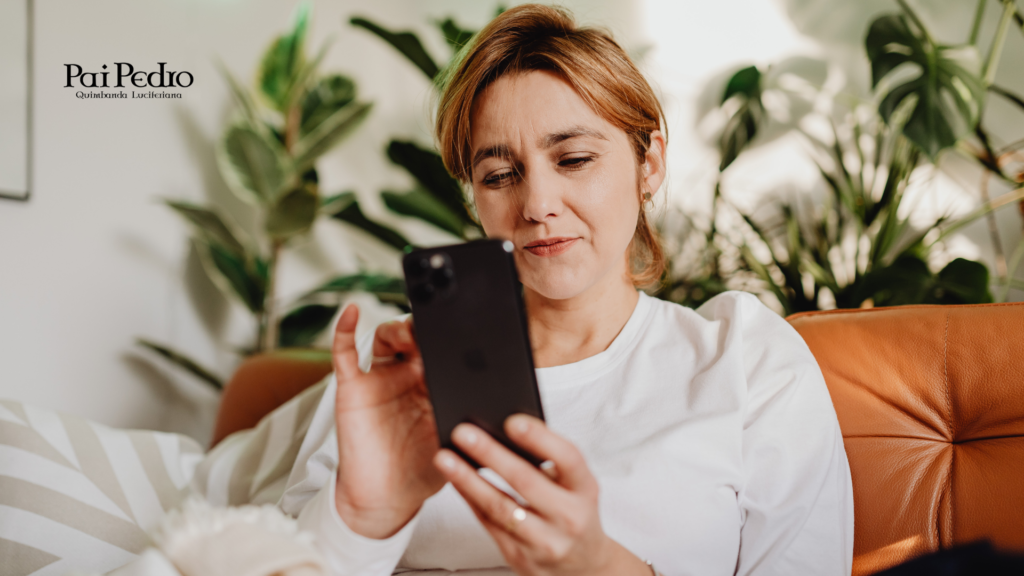 Mulher sentada no sofá olhando o celular com expressão pensativa, refletindo sobre mandar mensagem primeiro ou esperar uma resposta no relacionamento.